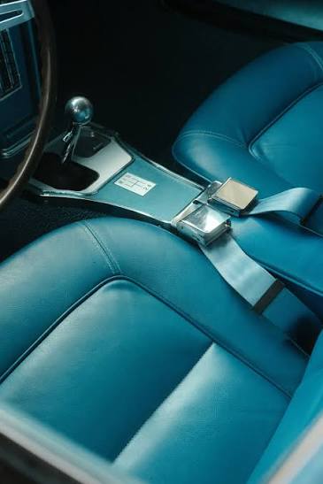 A vintage car interior in blue vinyl with a chrome-buckled lap belt lying across the seat beside the gear lever.