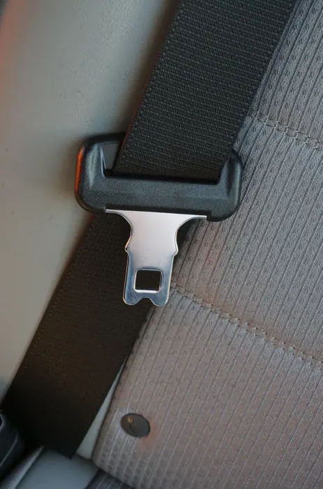 A seatbelt latch plate and webbing resting against a light grey seat, with the metal tongue and its slot clearly visible.