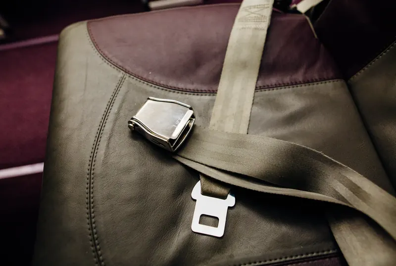 A lap-type seatbelt with a metal buckle lying across a leather seat, the webbing fed through a flat latch plate.