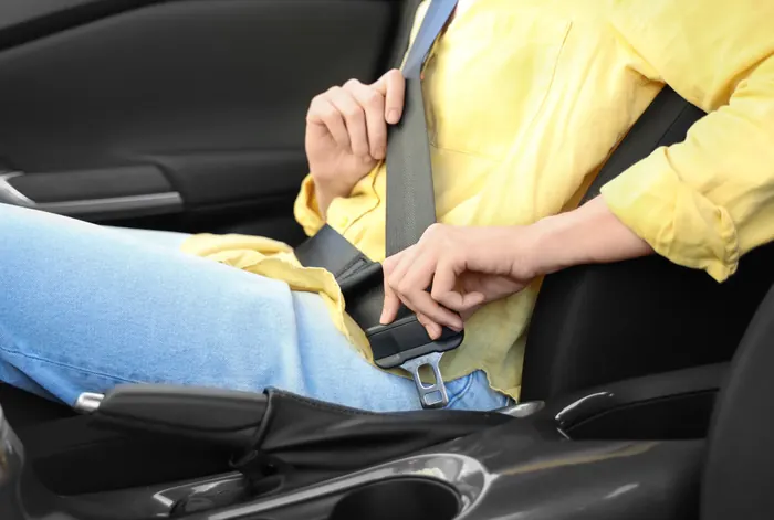 A person in a yellow shirt drawing a seatbelt across their lap and guiding the latch plate towards the buckle, seen from the side.